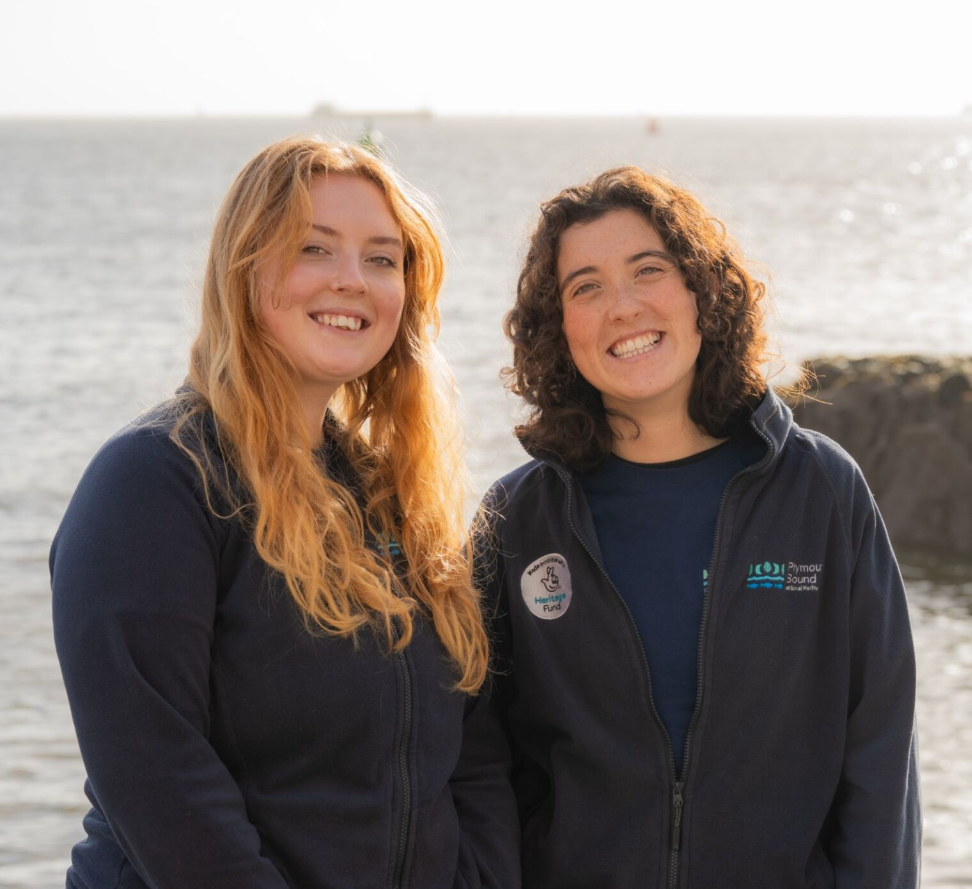 A photo of Plymouth Sound National Marine Park Coastal Rangers. On the left is Millie and on the Right Lilah. Plymouth Sound is the background.
