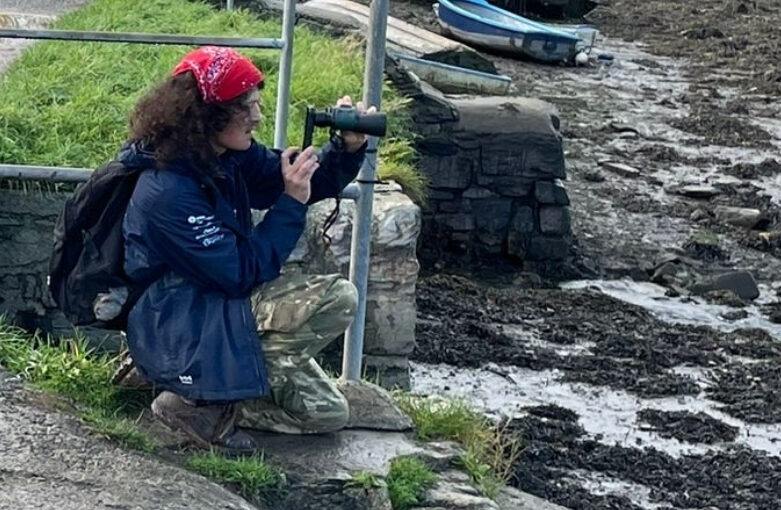 Coastal Ranger Luke, with curly black hair and a red bandana holding a pair of binoculars to his eyes, overlooking Ernesettle Creek for wading birds