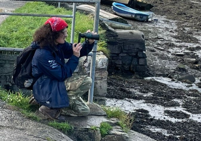 Coastal Ranger Luke, with curly black hair and a red bandana holding a pair of binoculars to his eyes, overlooking Ernesettle Creek for wading birds