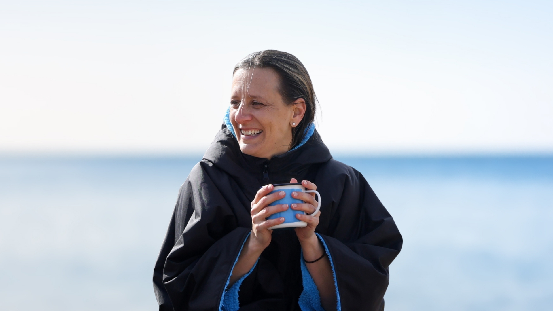 Woman on a beach in a Dry Robe and a mug of hot tea in her hands